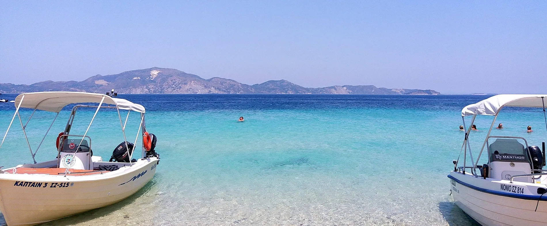 Two small motorboats docked on a clear turquoise beach with people swimming and mountainous islands in the distance.