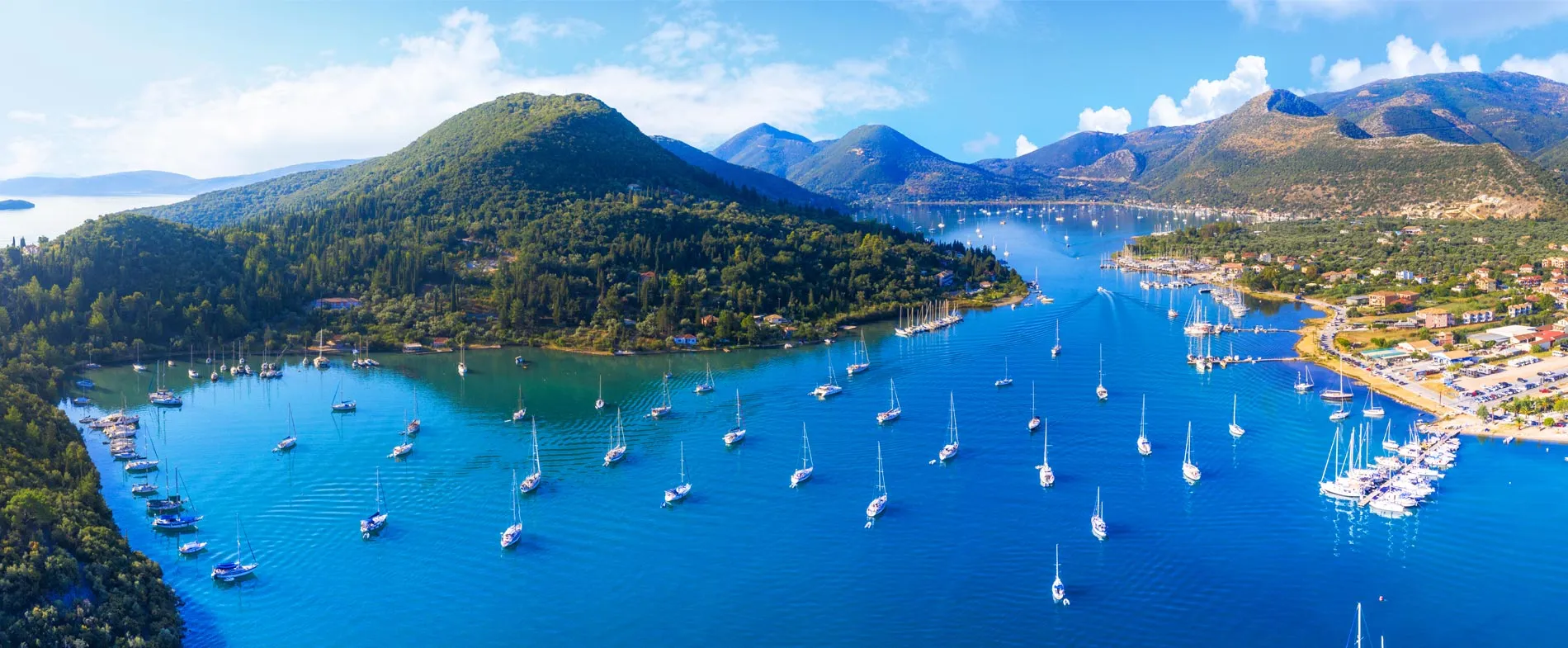 Bay view with numerous sailboats anchored near green hills and a coastal town under a bright sky.