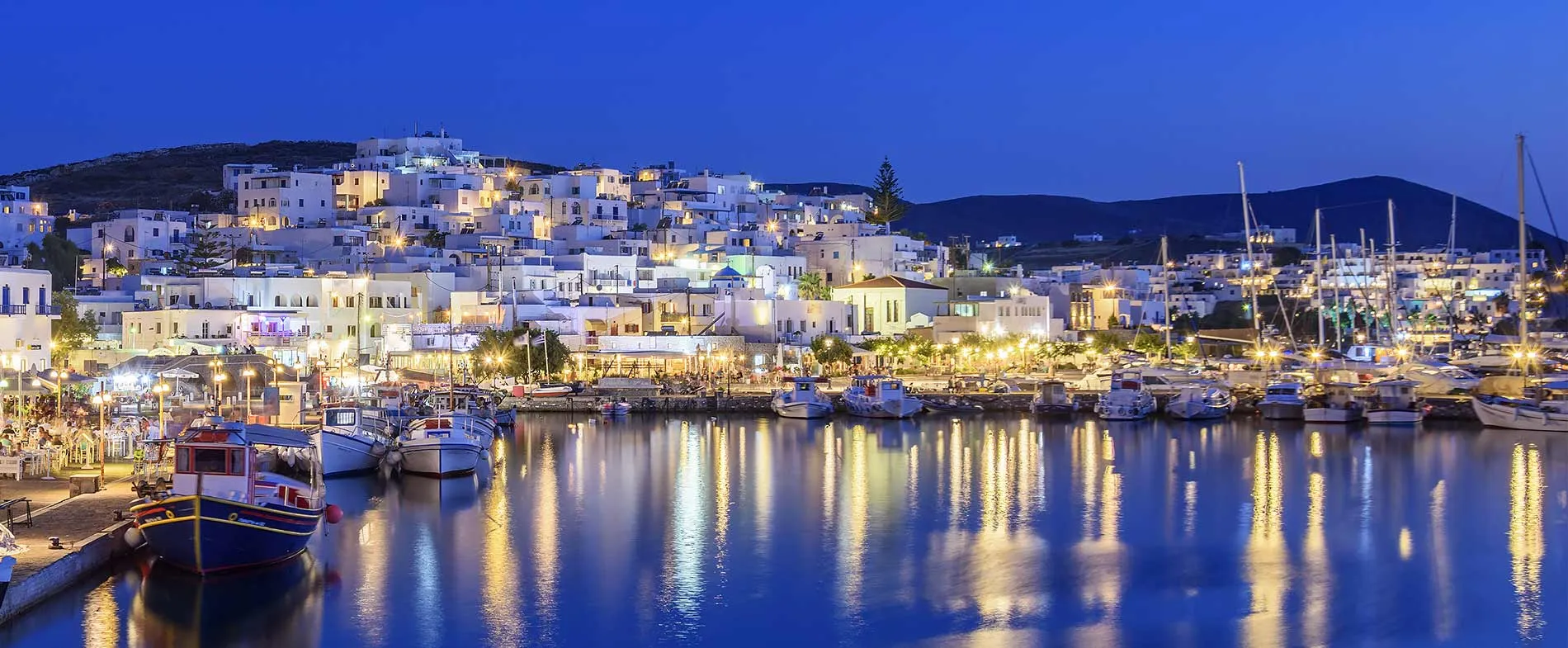 Porto di Naxos di notte con barche e case bianche illuminate lungo la riva.