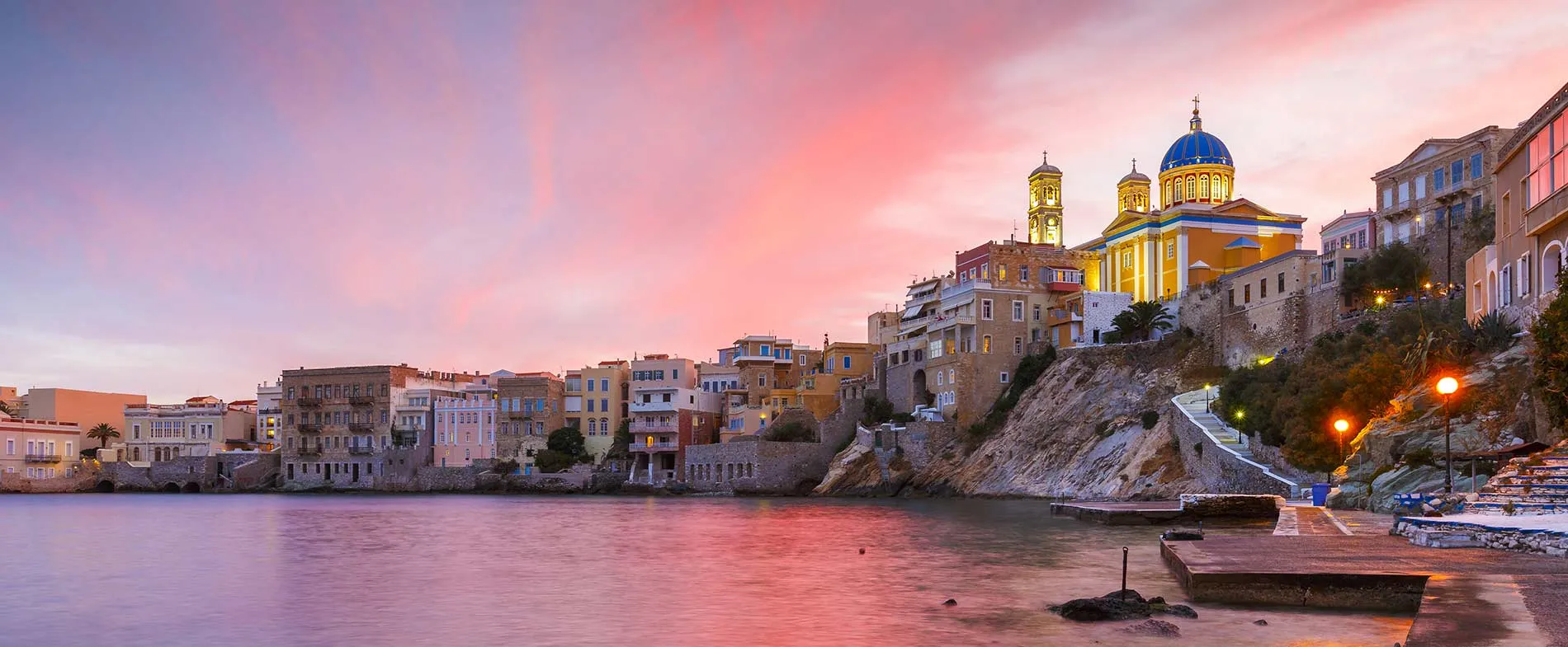 Lungomare dell'isola di Syros con edifici colorati e una chiesa al tramonto sotto un cielo rosa e viola.