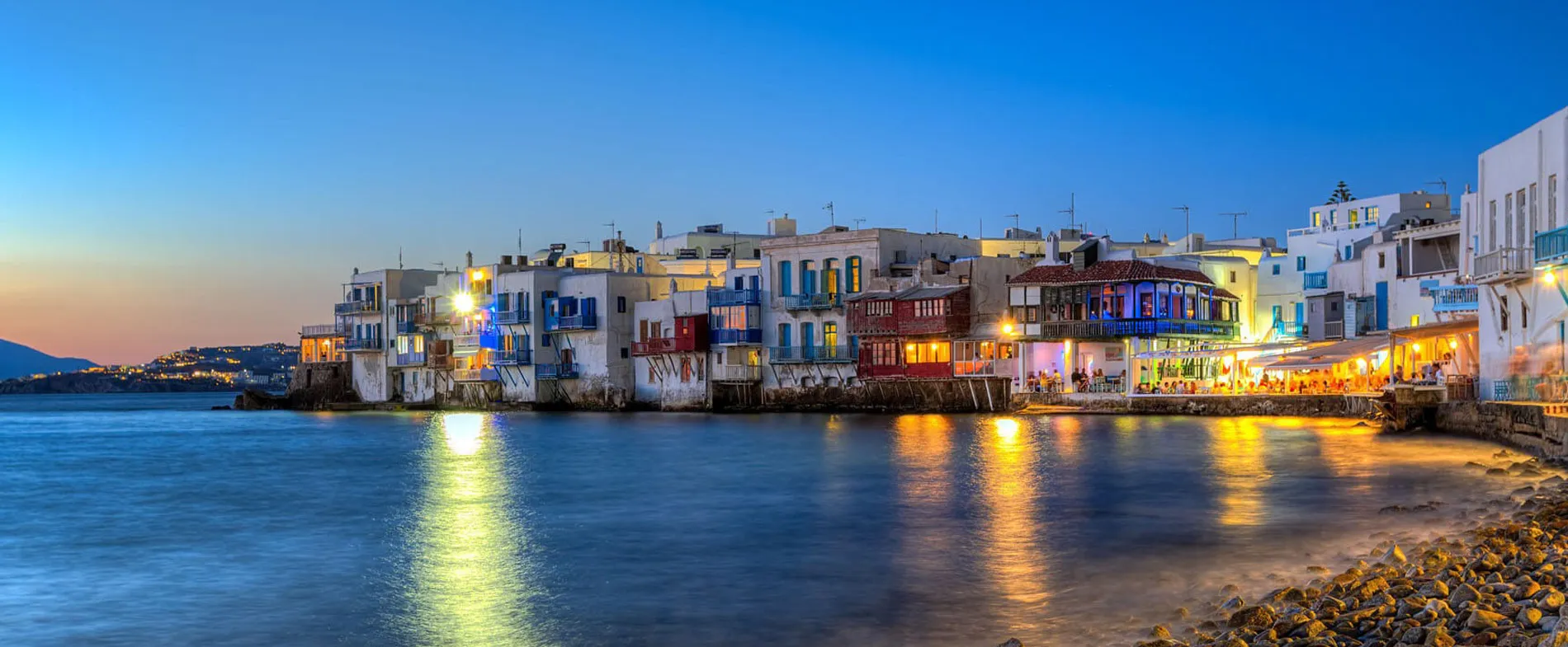 Lungomare di Mykonos con edifici bianchi tradizionali e mare calmo al tramonto per noleggio auto.