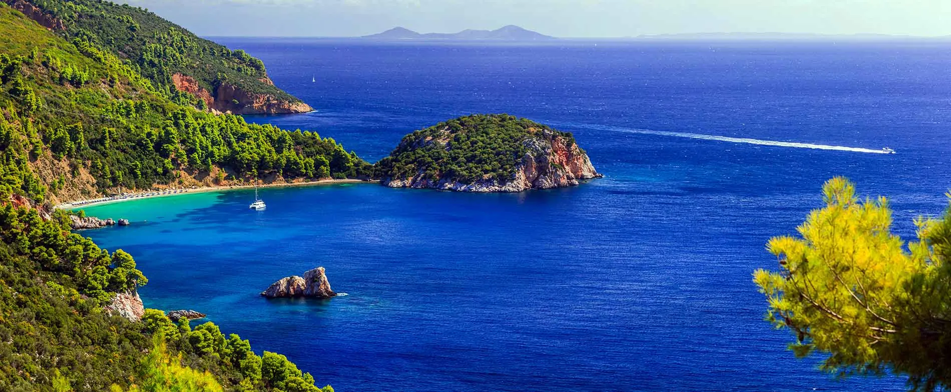 Havsvik med gröna klippor och segelbåt i klart blått vatten vid Skopelos, Grekland.
