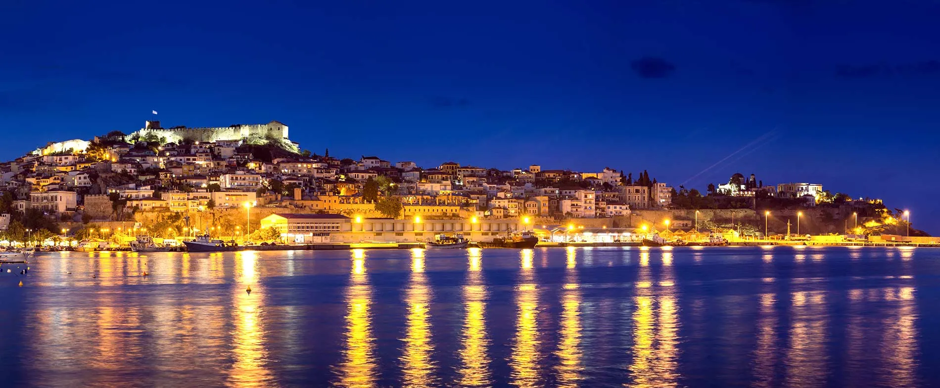 Vue nocturne du port de Kavala avec bâtiments illuminés et château reflétés sur l'eau calme pour location voiture.