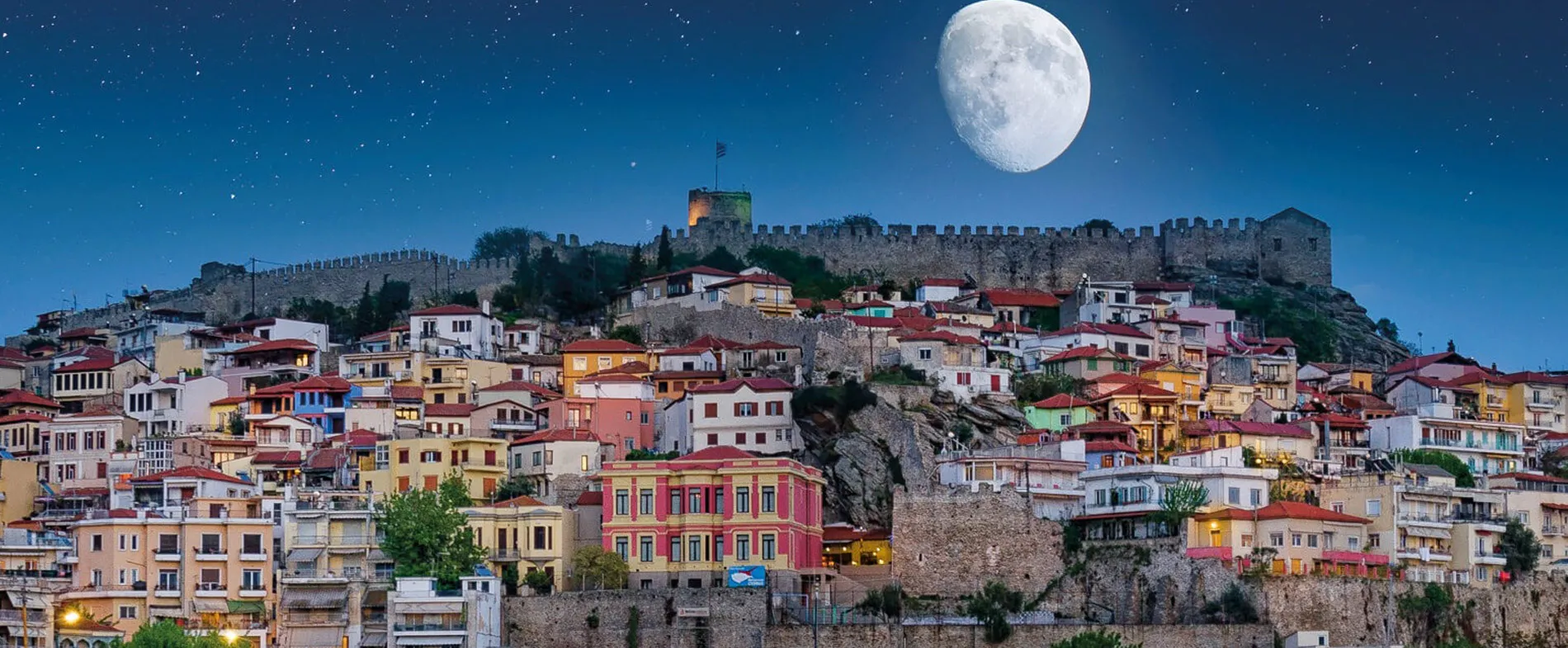 Veduta notturna di Kavala con castello, cielo stellato e luna grande visibile.