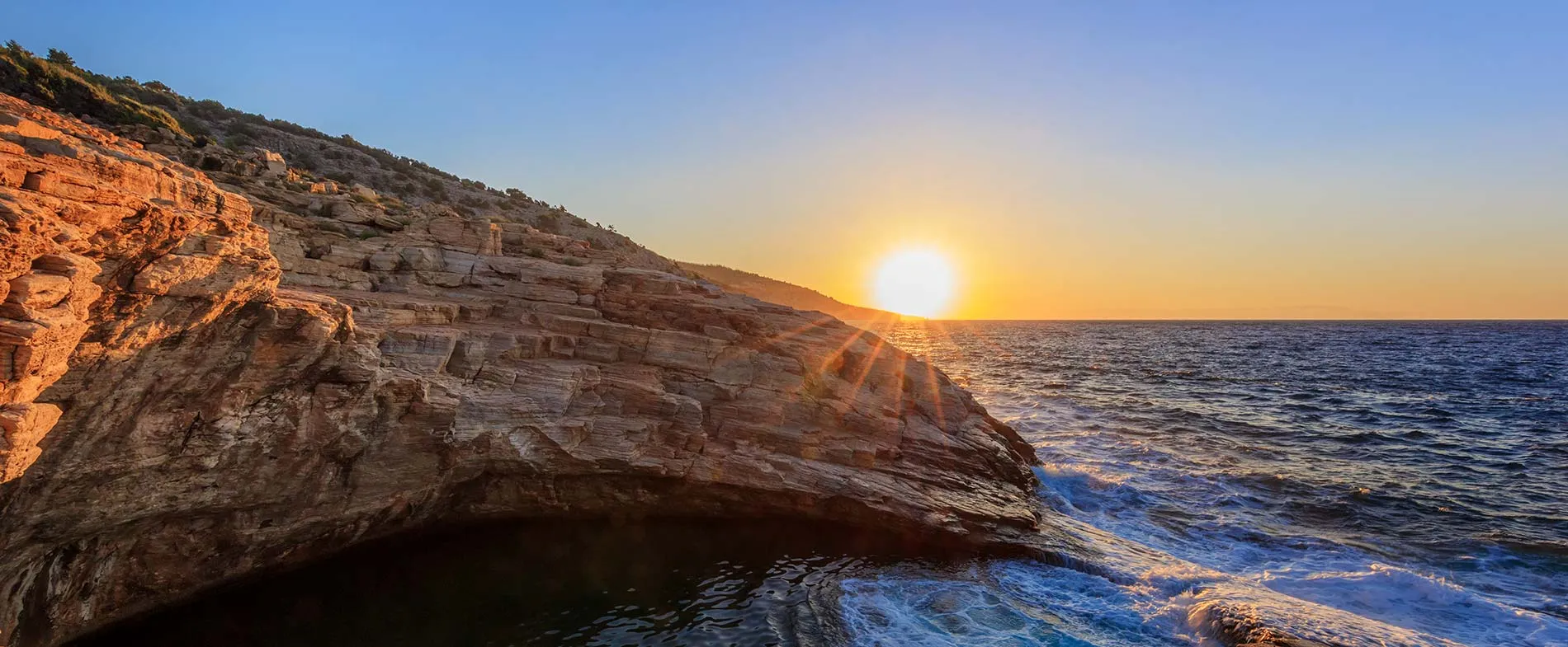 Costa rocciosa al tramonto con onde che si infrangono sulle scogliere sotto un cielo limpido.