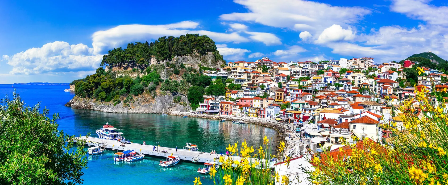 Costa di Parga con porto, case colorate e barche su acqua limpida sotto un cielo parzialmente nuvoloso.