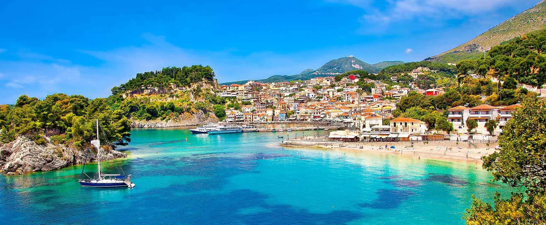 Parga seaside town with turquoise waters, boats, and hillside houses under a bright blue sky.