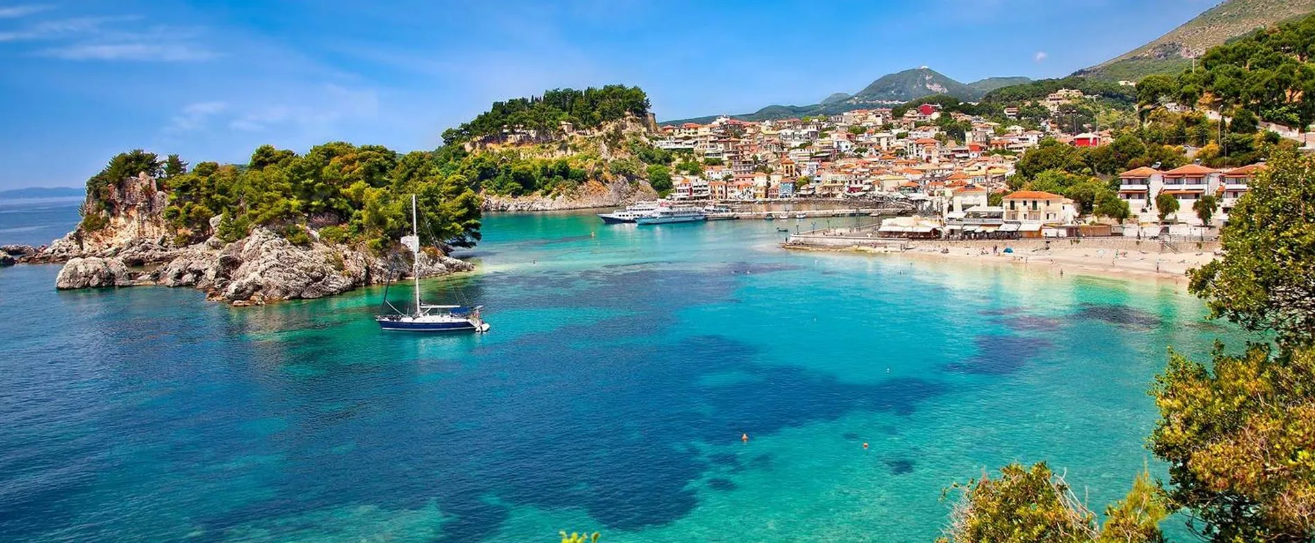 Costa di Parga con mare turchese, barche e città sulla collina sotto un cielo blu limpido.