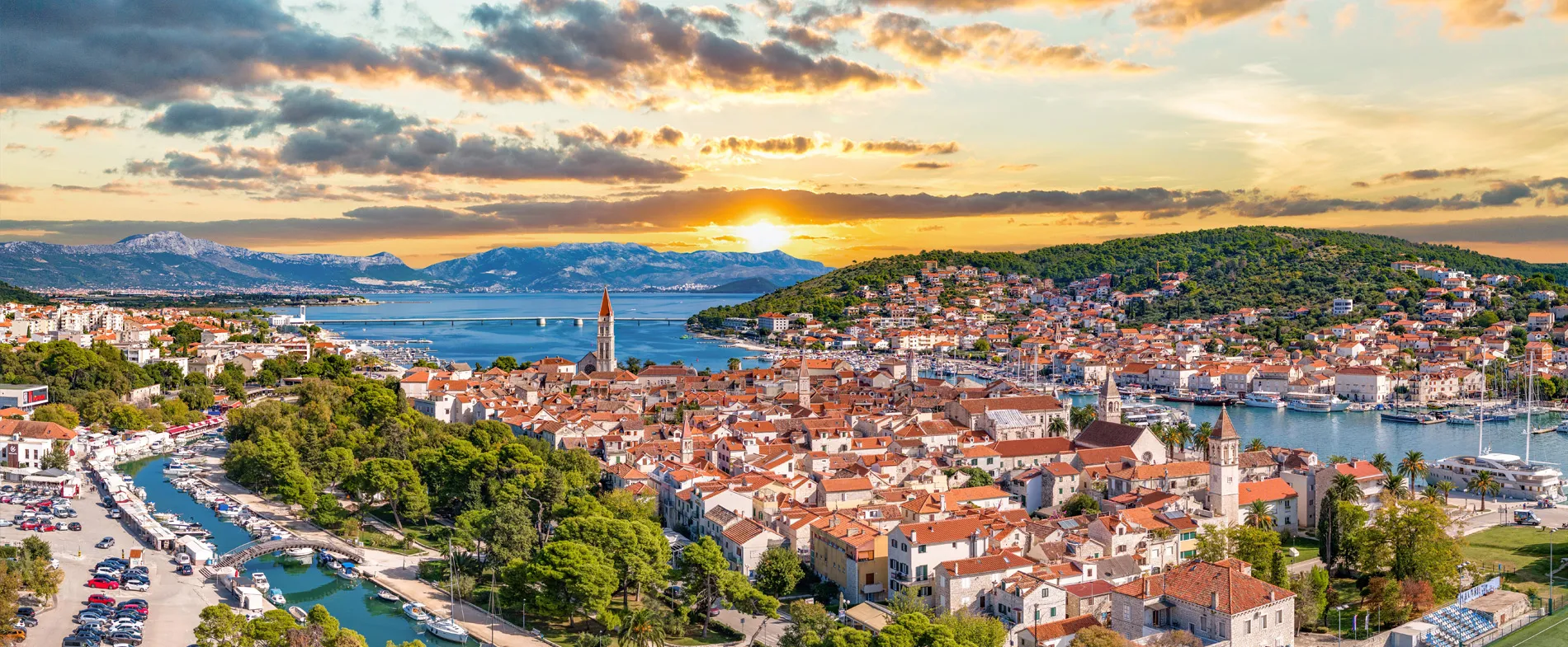 Trogir town with red-roofed buildings near a marina and mountains during sunset in Croatia.