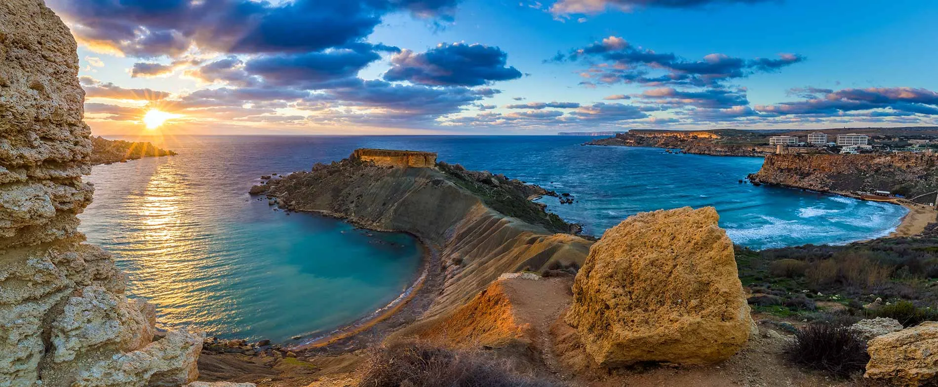 Golden sunset over the rugged cliffs and turquoise bay at Golden Bay, Malta, with calm sea and scattered clouds.