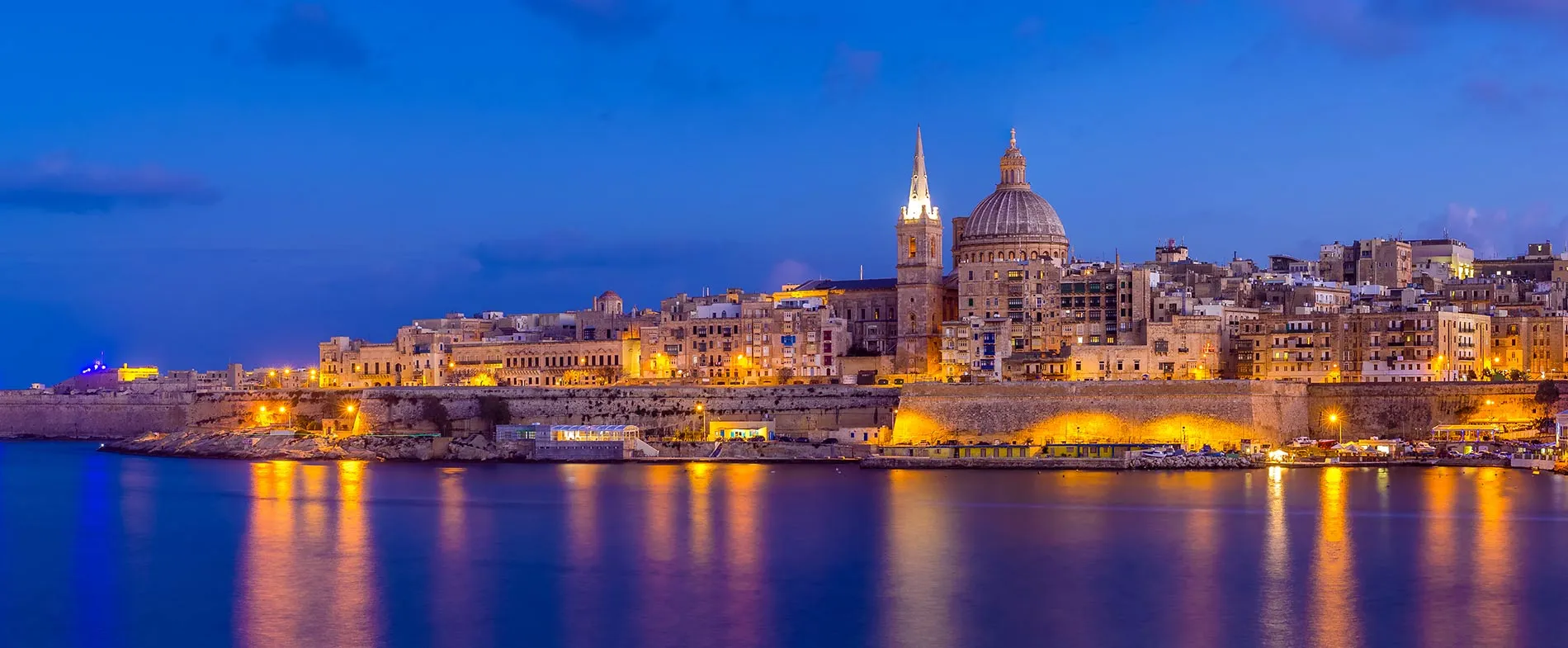 Malta skyline with historic buildings and the dome of St. Paul's Cathedral lit warmly at dusk by the water.