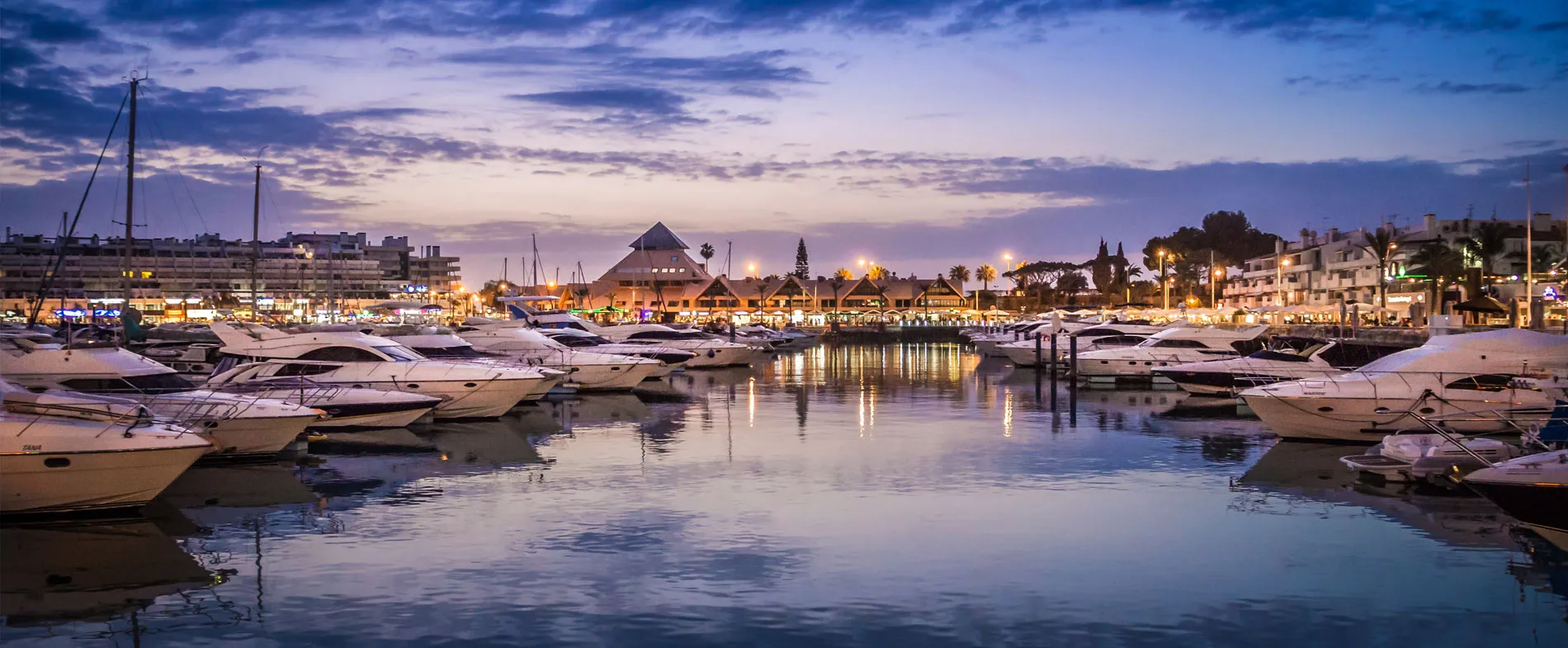 Puerto Banús marina at dusk with luxury boats docked and illuminated waterfront buildings.