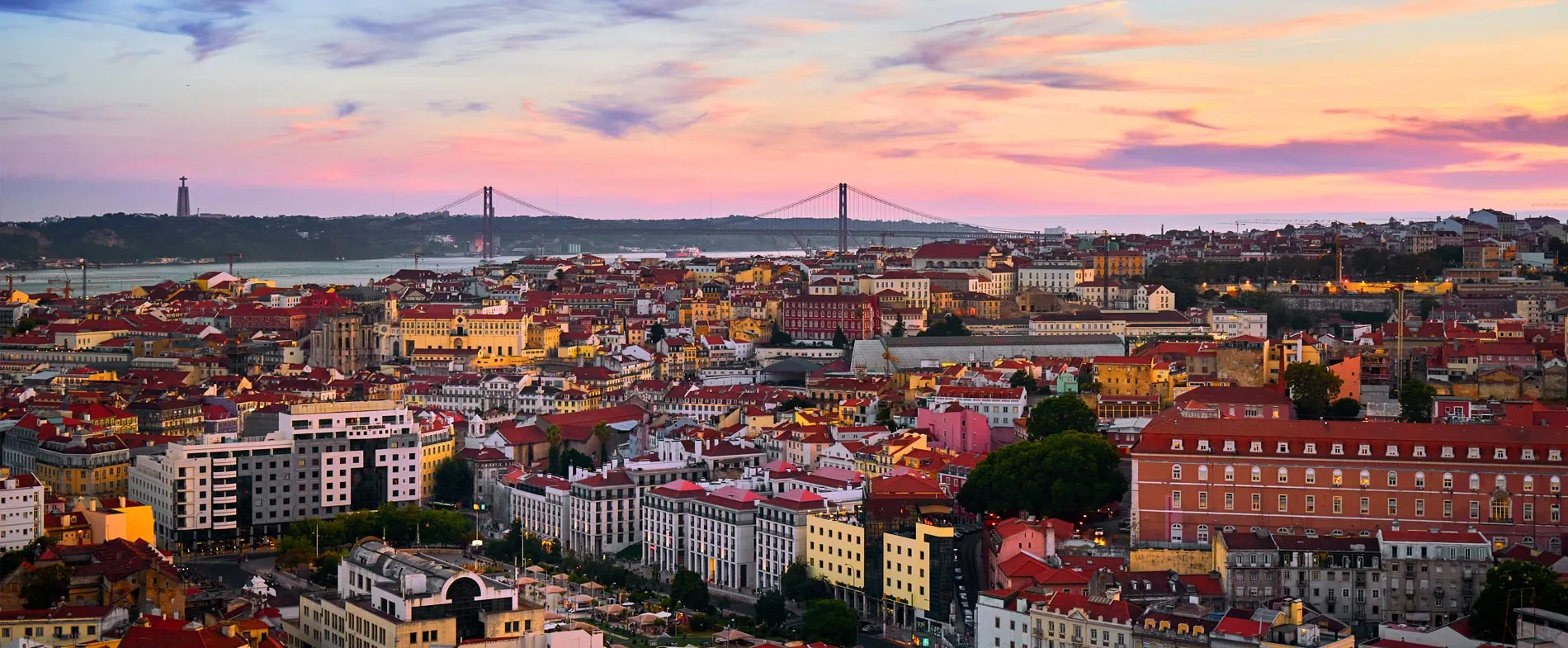 Lisbon cityscape at sunset with red rooftops and the 25 de Abril Bridge over the Tagus River.