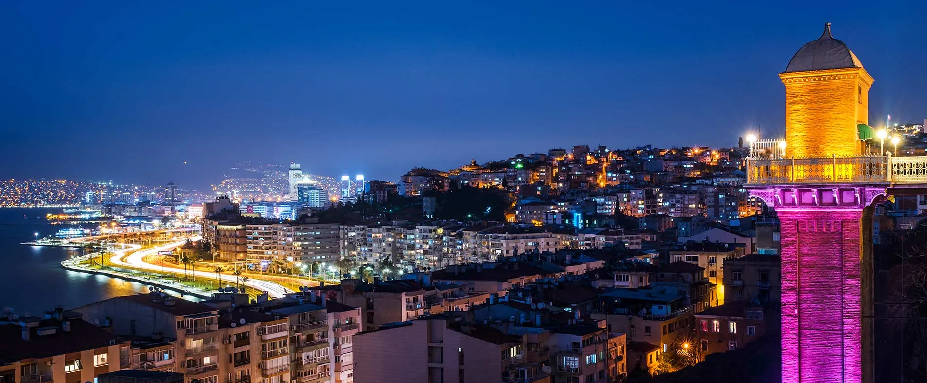 İzmir, Türkiye gece manzarası, kıyı boyunca aydınlatılmış sokaklar ve mor ile altın renkli tarihi asansör kulesi.