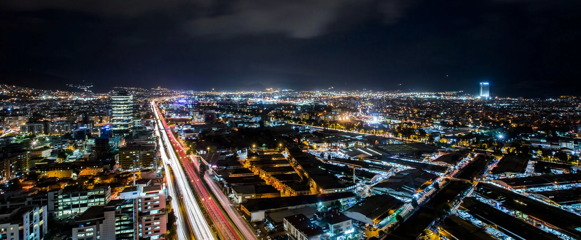 Karanlık gökyüzü altında yoğun ışıklı otoyol ve binalarla gece şehir manzarası.