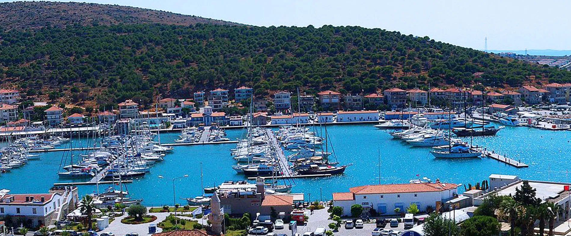 Marina, temiz mavi sularda demirlemiş tekneler ve arka planda yeşil tepeler.