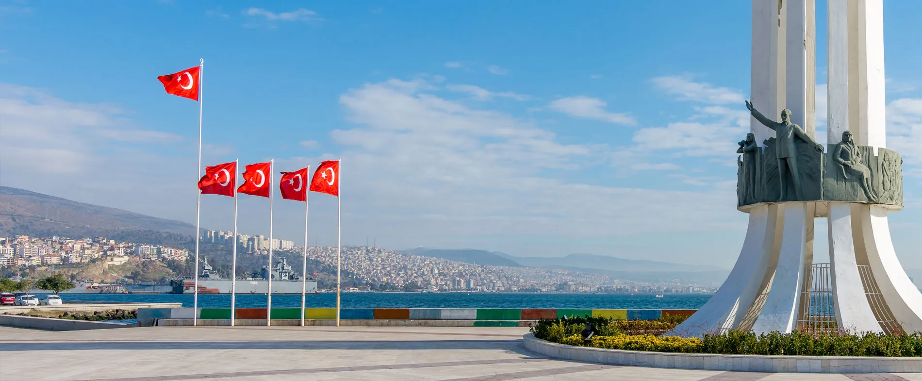 İzmir sahilinde Türk bayrakları ve su kenarında büyük bir heykel, güneşli bir günde.