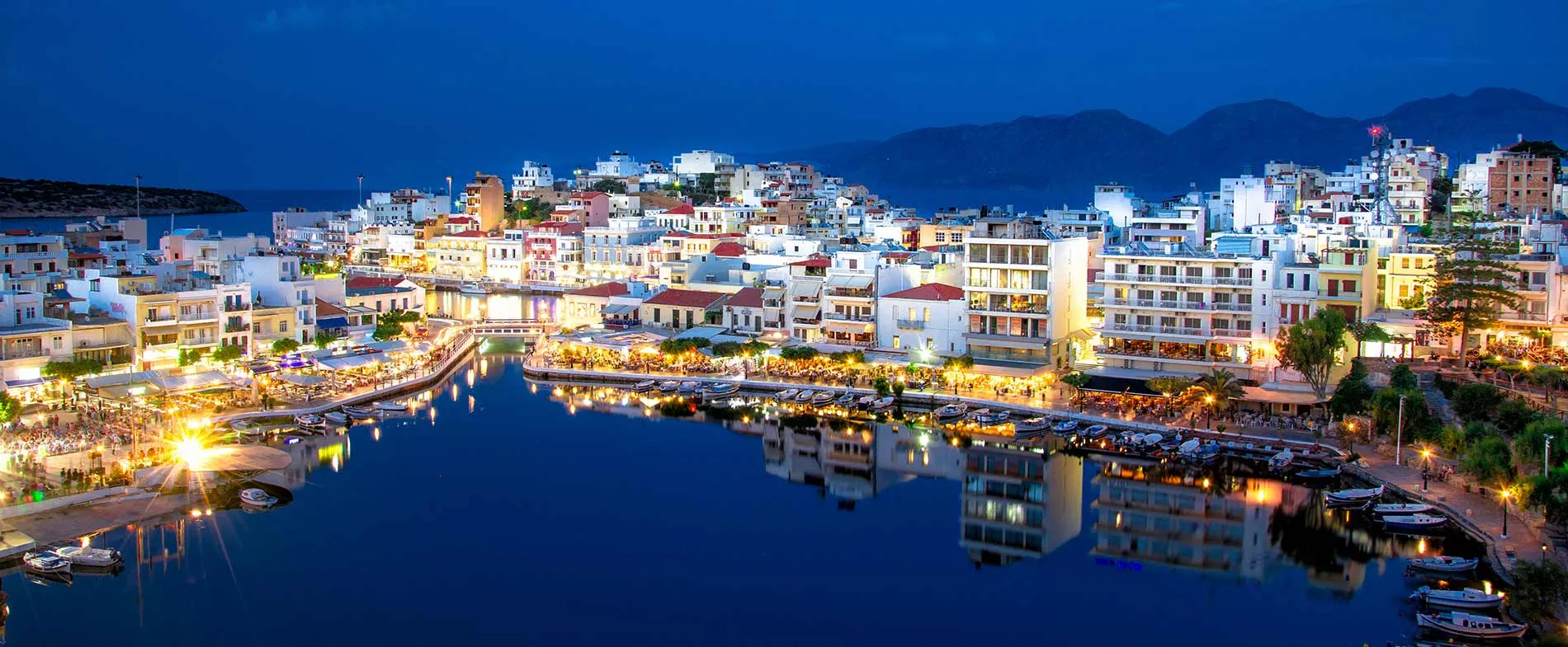Ville d'Agios Nikolaos la nuit avec bâtiments illuminés et reflets dans le port calme.