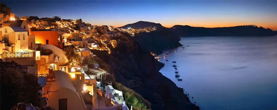 Santorini coastline at night with illuminated houses and boats on calm sea waters.