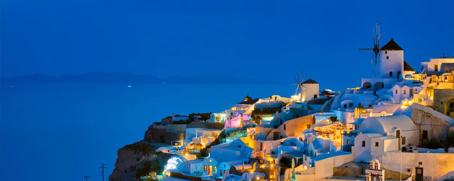 Santorini coastline with illuminated buildings and traditional windmills at dusk for car rental.