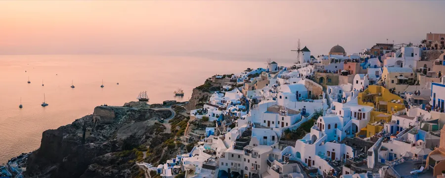 Santorini coastline at sunset with white buildings and sailboats on the calm sea for car rental.