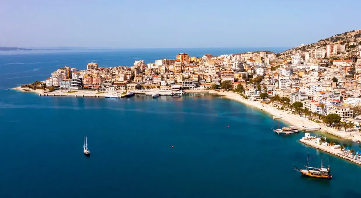 Saranda coastline with buildings and boats along the shore under a clear sky.