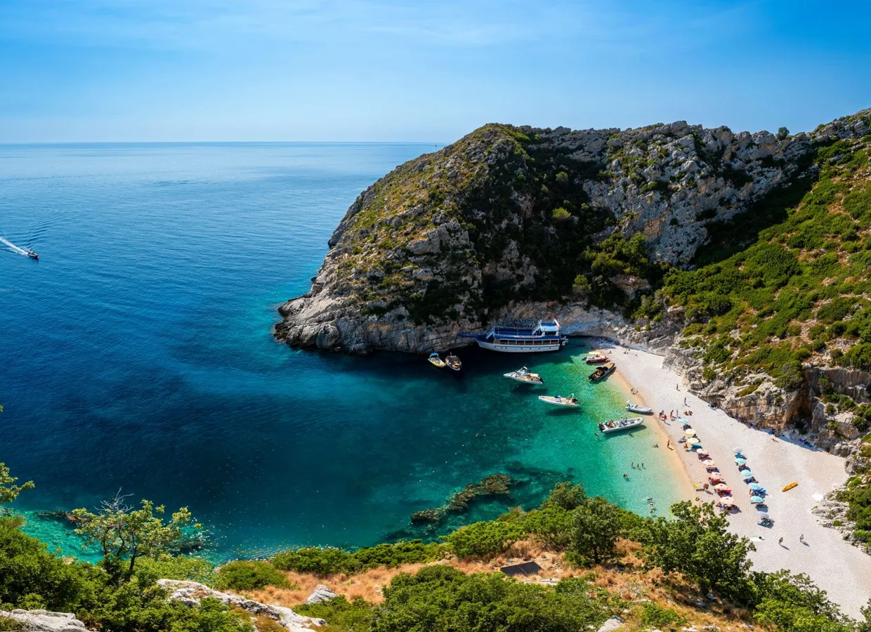 Secluded beach cove with clear turquoise water, boats anchored, and umbrellas along the shore.