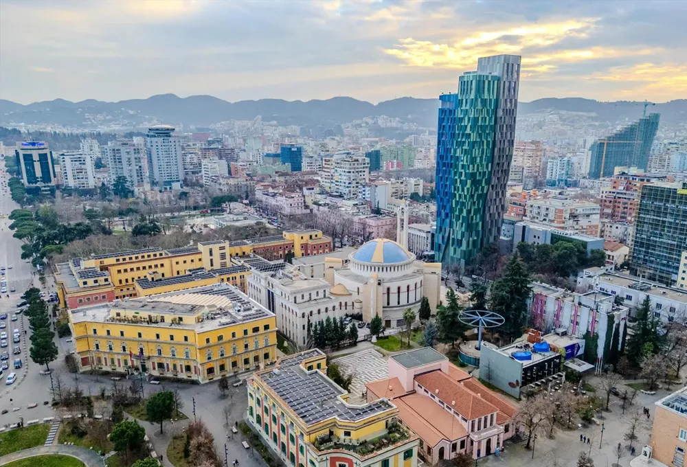 Tirana city skyline with modern and historic buildings under a cloudy sky at sunset.