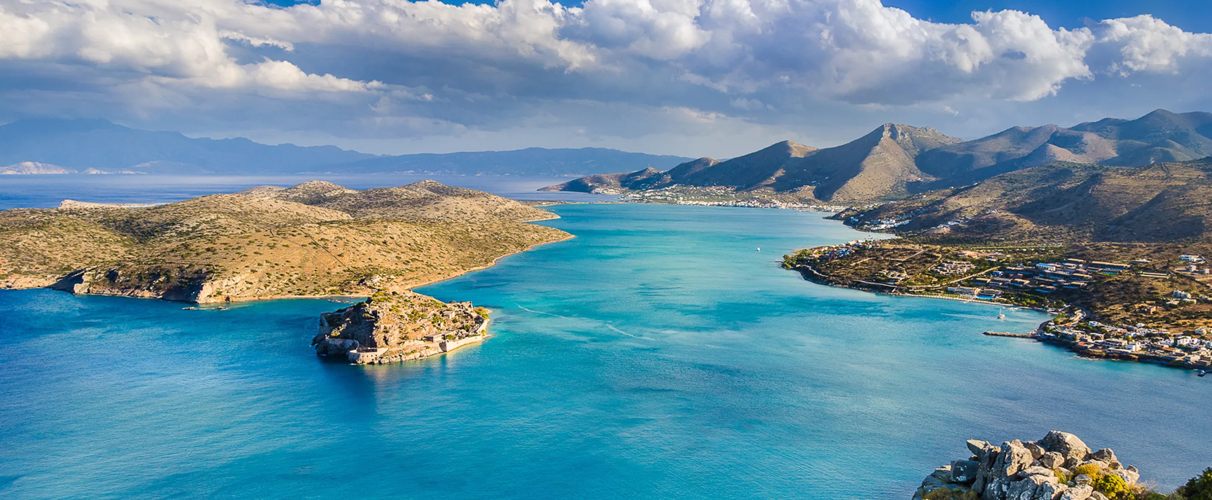 Elounda Bay coastline with clear blue water and mountainous landscape under a partly cloudy sky.