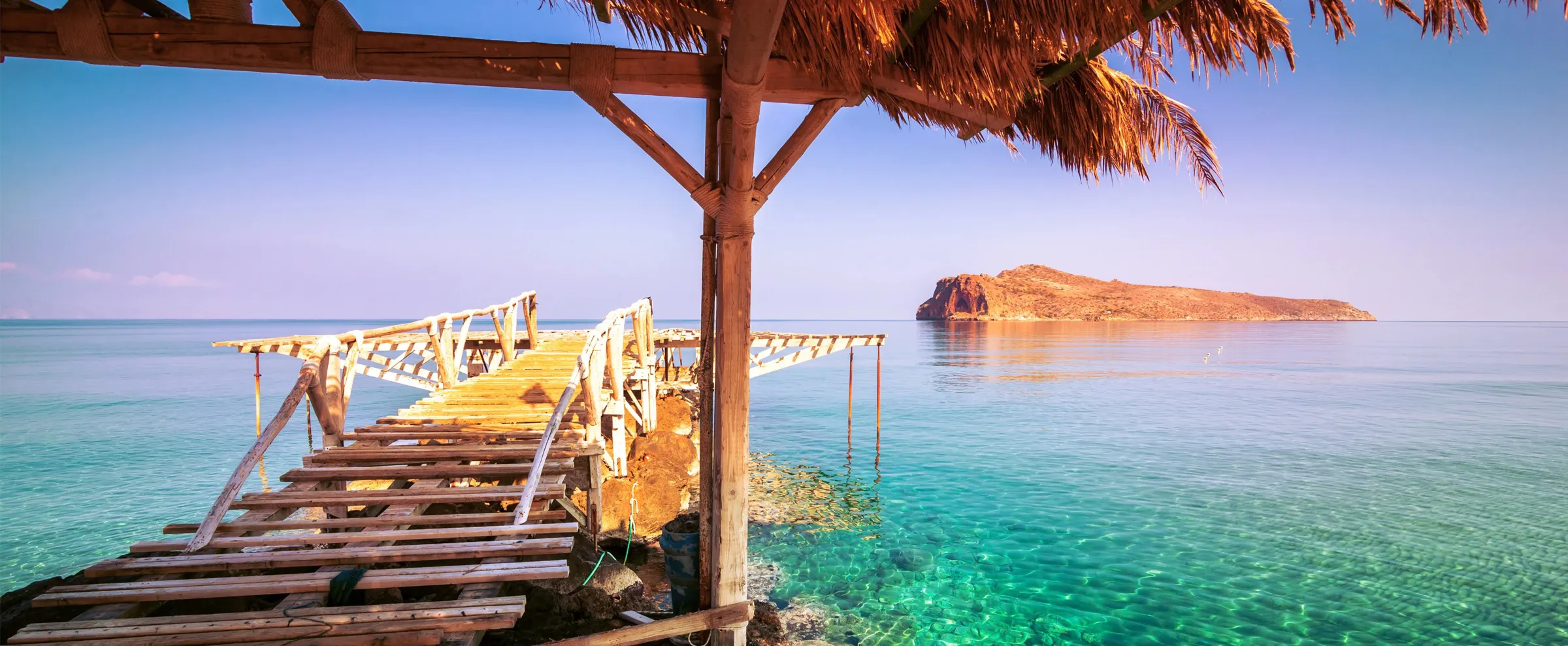 Wooden pier with a thatched roof over clear turquoise sea water and a rocky island in the distance.