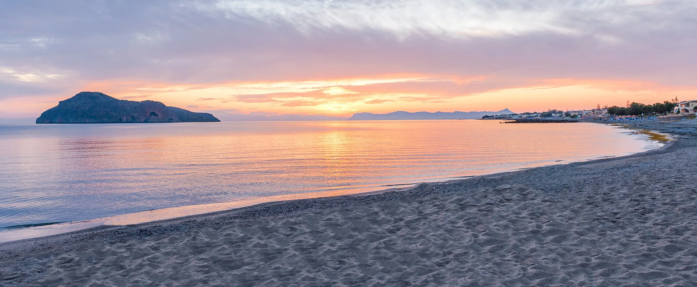 Sunset over a calm beach with an island and distant mountains in the background for a peaceful travel scene.