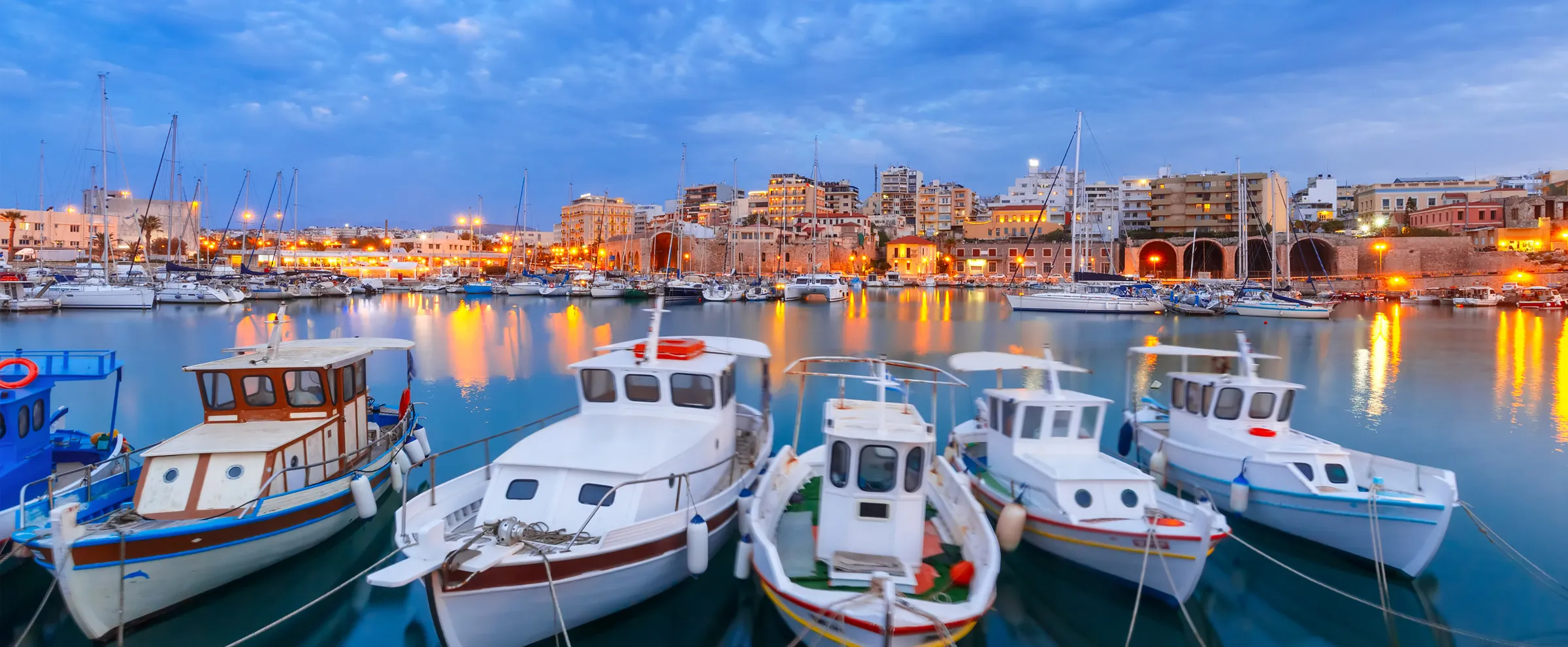 Porto con barche ormeggiate a Heraklion, Creta, Grecia, sotto un cielo blu al crepuscolo con luci cittadine.