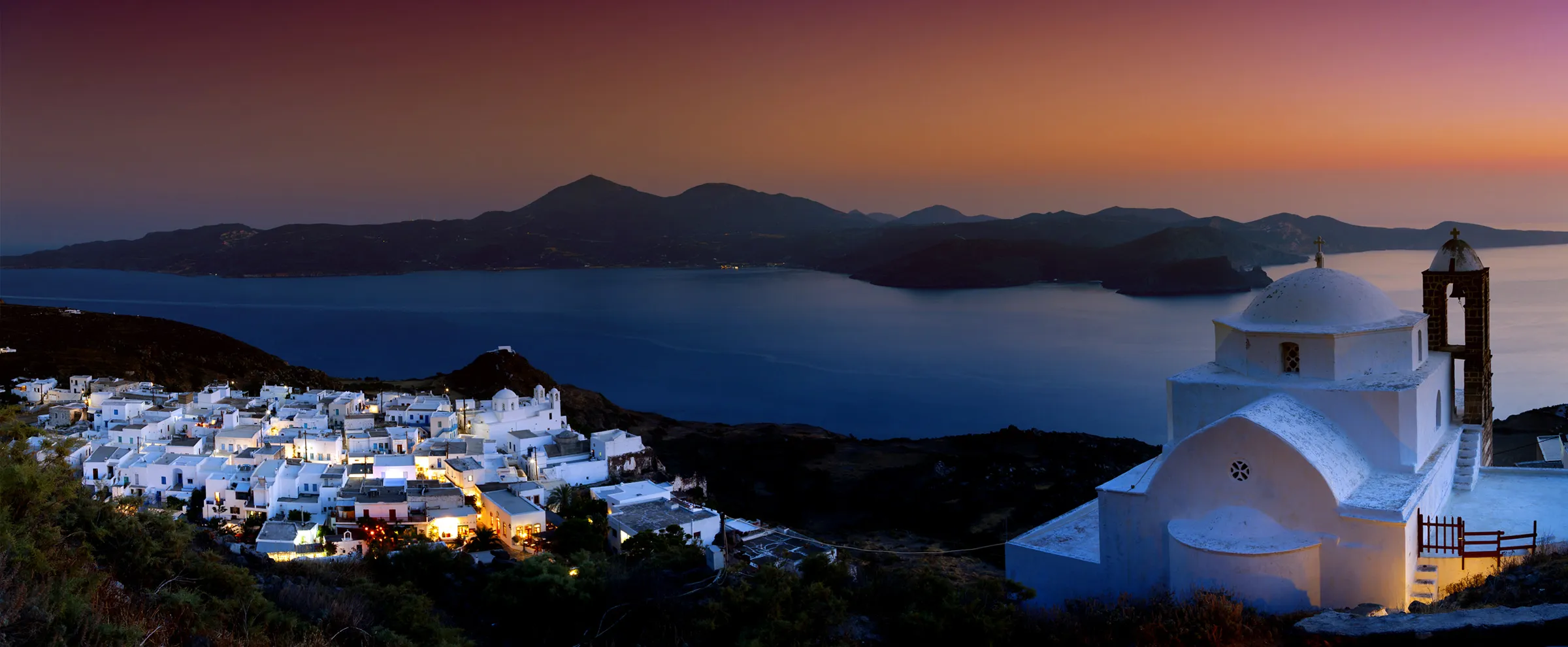 Villaggio dell'isola greca al tramonto con chiesa bianca e vista sul mare calmo e sulle montagne lontane.