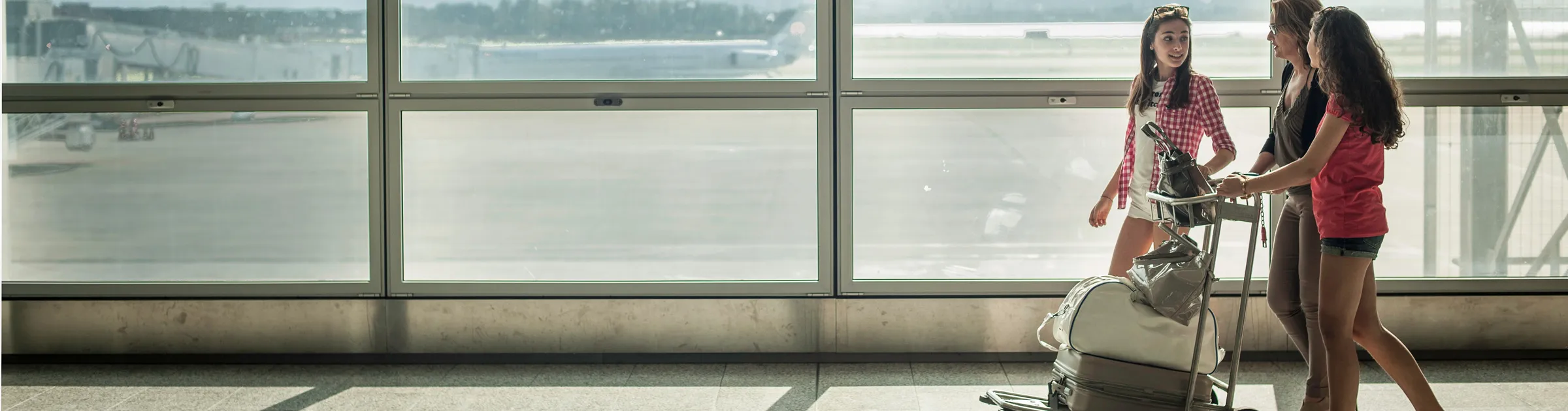 Three women walking with luggage trolley inside an airport terminal near large windows.