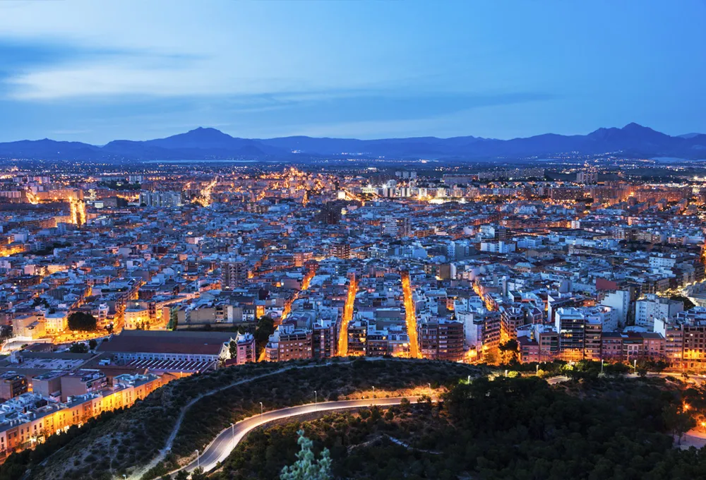 Vedere de noapte în Murcia cu străzi luminate și zonă urbană cu munți în fundal.