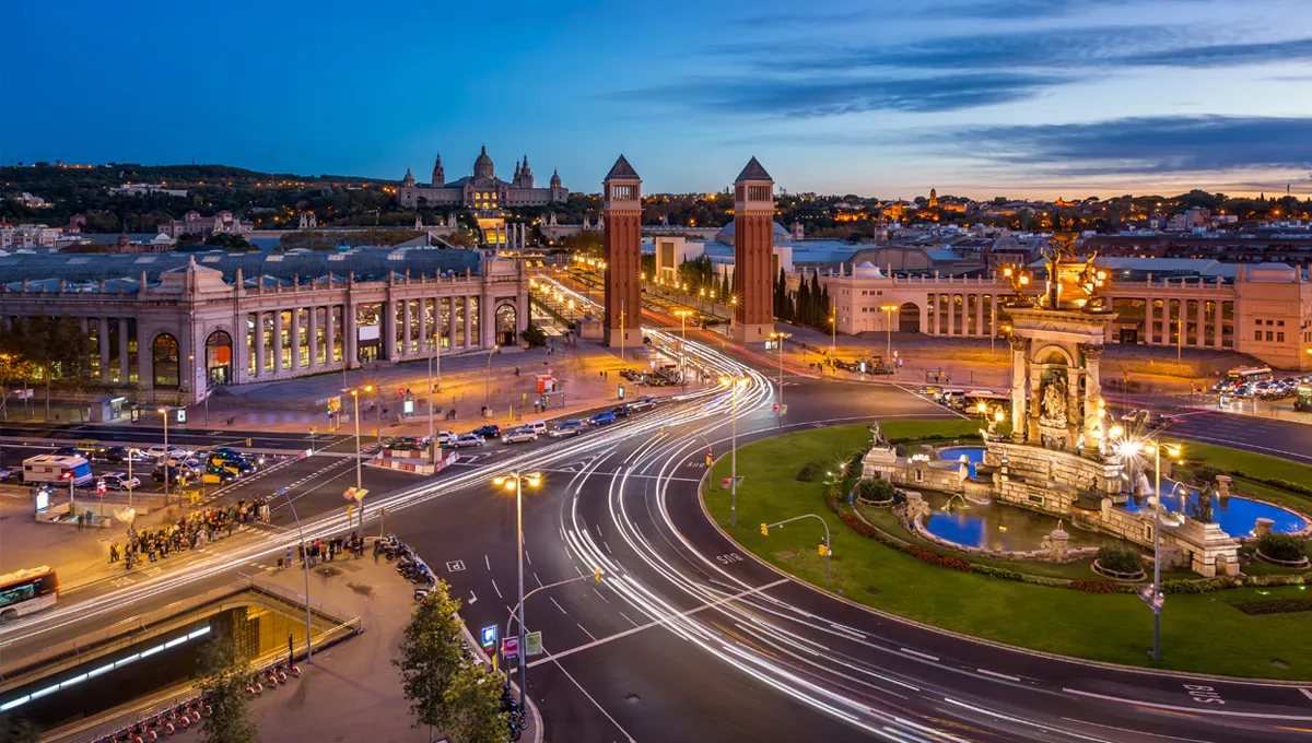 Piața Spaniei din Barcelona la amurg cu urme luminoase de mașini în sensul giratoriu lângă turnuri renumite.