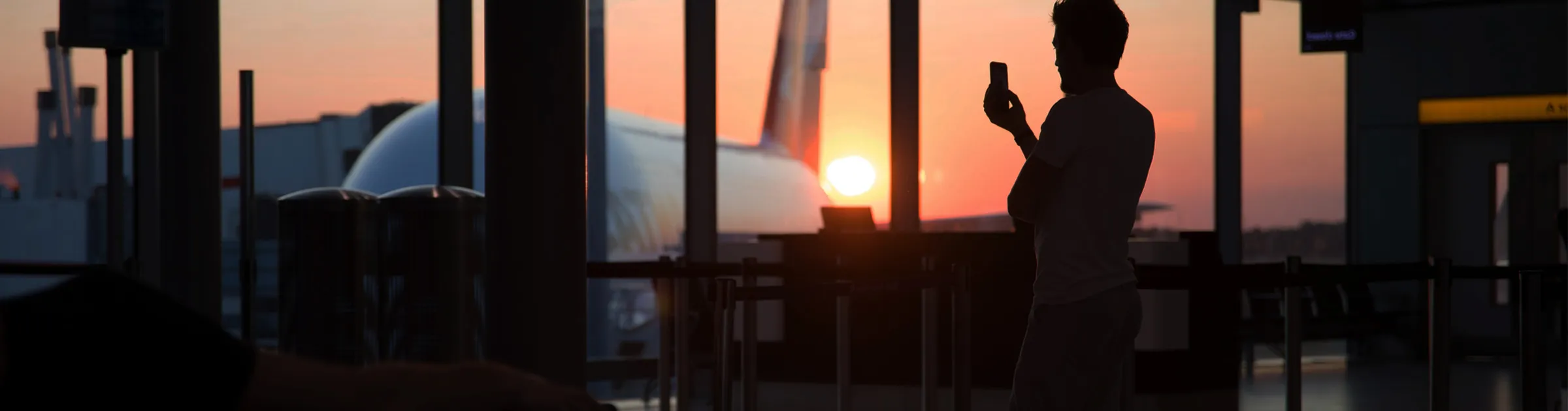 Sunset at airport terminal with silhouette of person taking photo of airplane outside window.