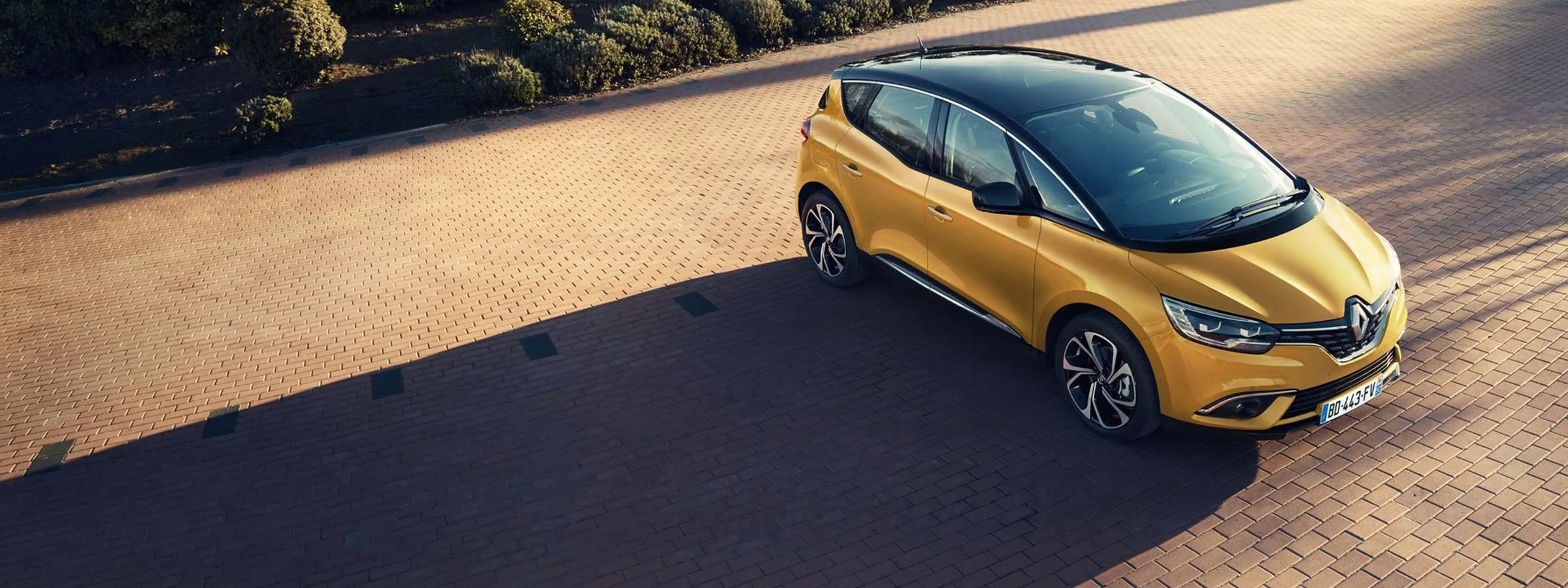 Mașină compactă Renault de închiriat, galbenă cu acoperiș negru, parcată pe o alee din cărămidă.