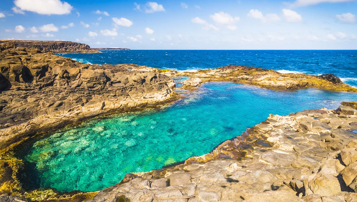 Piscină naturală de rocă vulcanică cu apă turcoaz limpede lângă ocean sub un cer parțial noros.