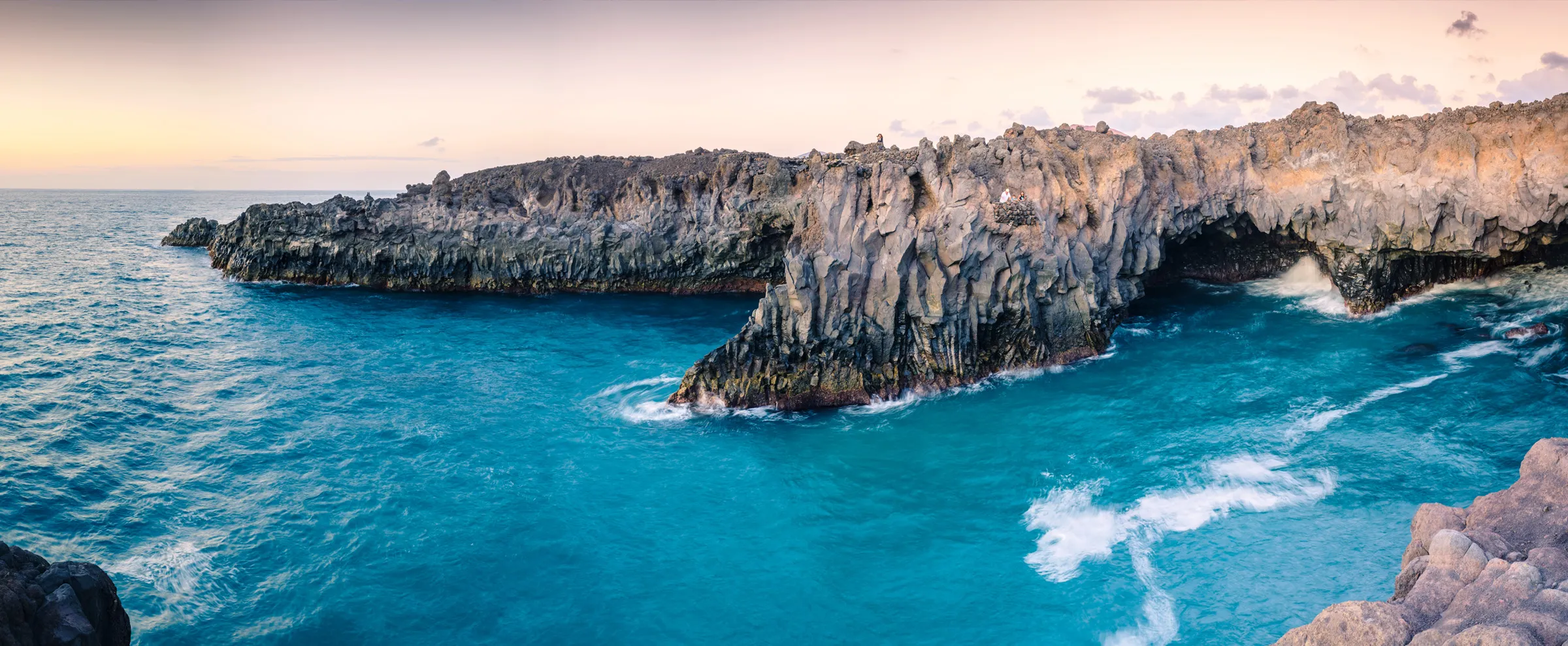Natürliche felsige Küstenlinie mit blauen Wellen, die bei ruhigem Sonnenuntergang auf die Felsen schlagen.