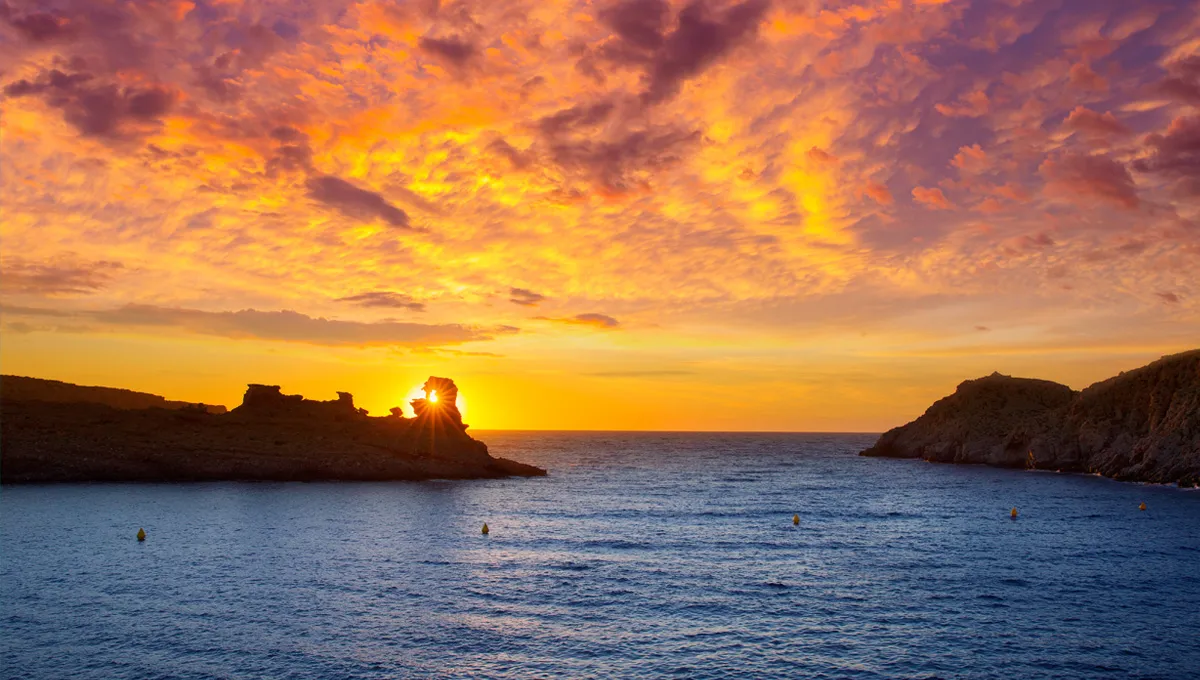 Tramonto su costa rocciosa con nuvole colorate e mare calmo.