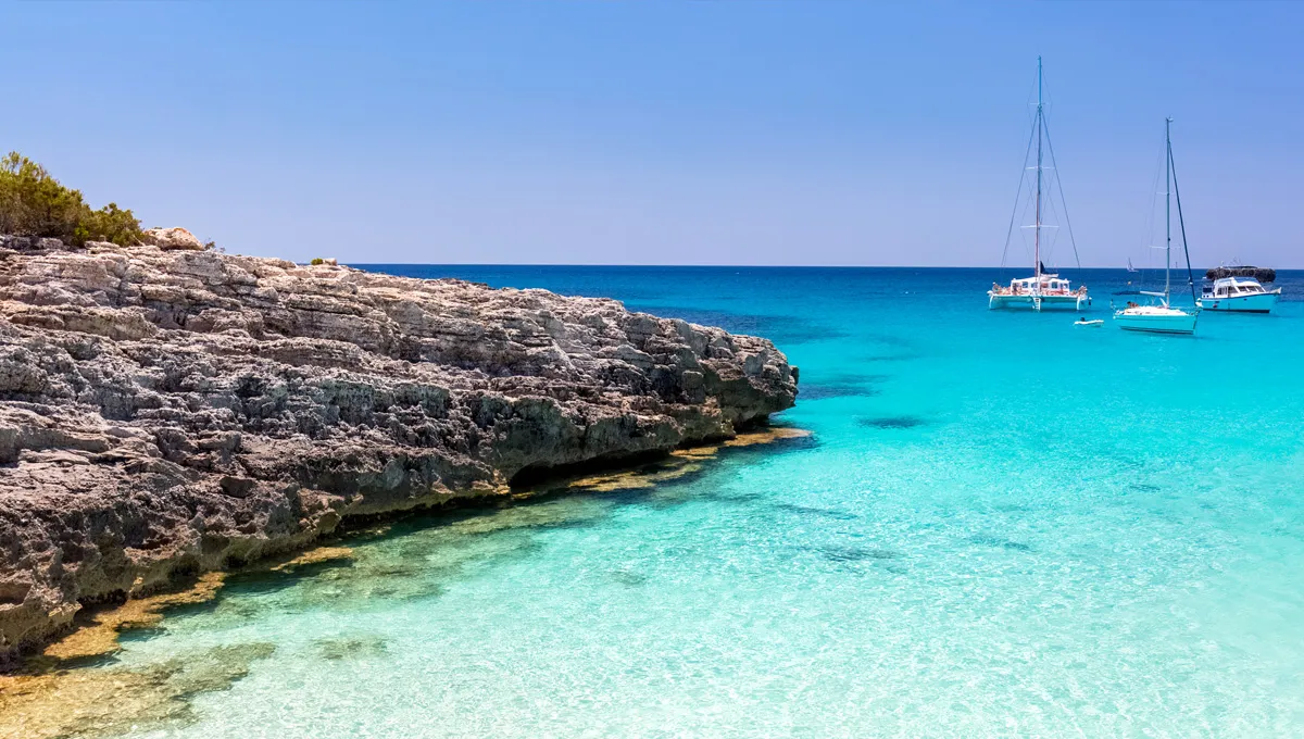 Acque turchesi costiere con barche a vela e formazioni rocciose in una giornata soleggiata in una cala mediterranea.