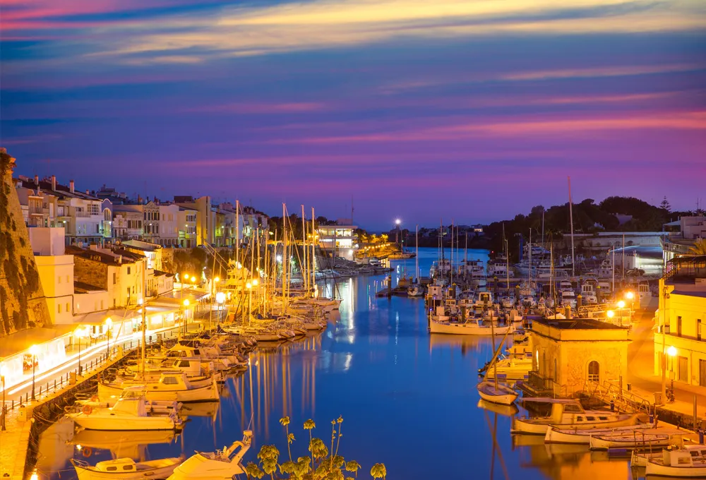 Porto a Ciutadella de Menorca con yacht e barche attraccati al tramonto e luci stradali accese.