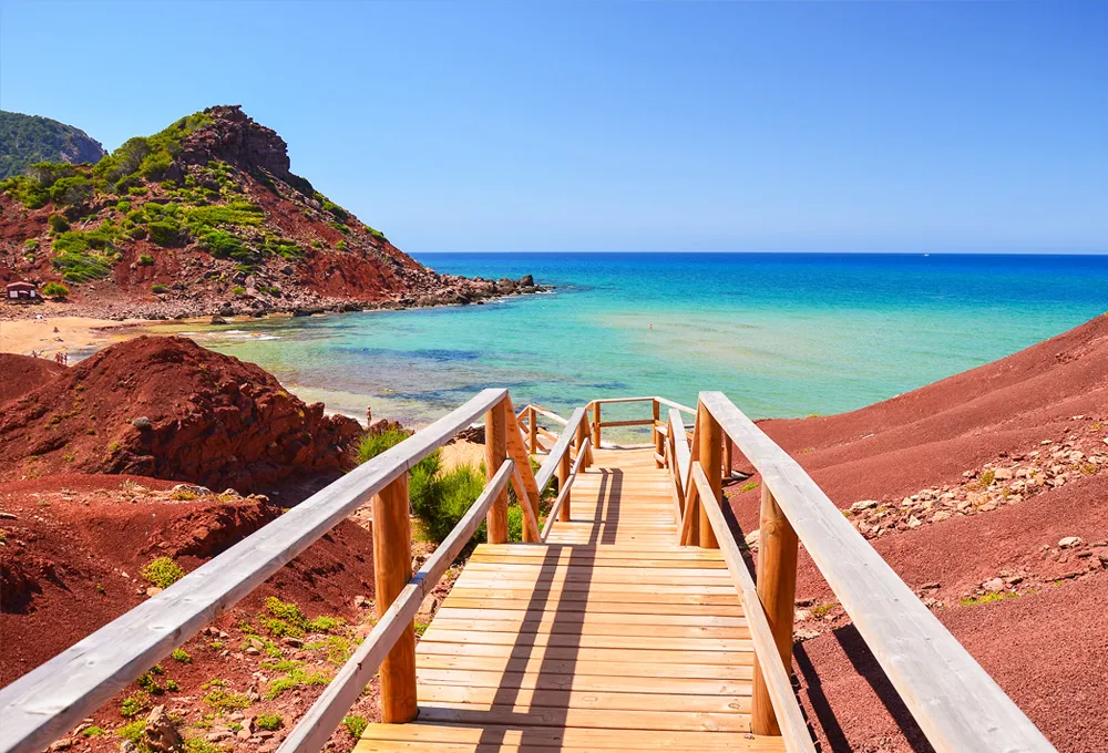 Passerella in legno che conduce a una spiaggia rocciosa rossa con acqua turchese chiara e cielo azzurro.