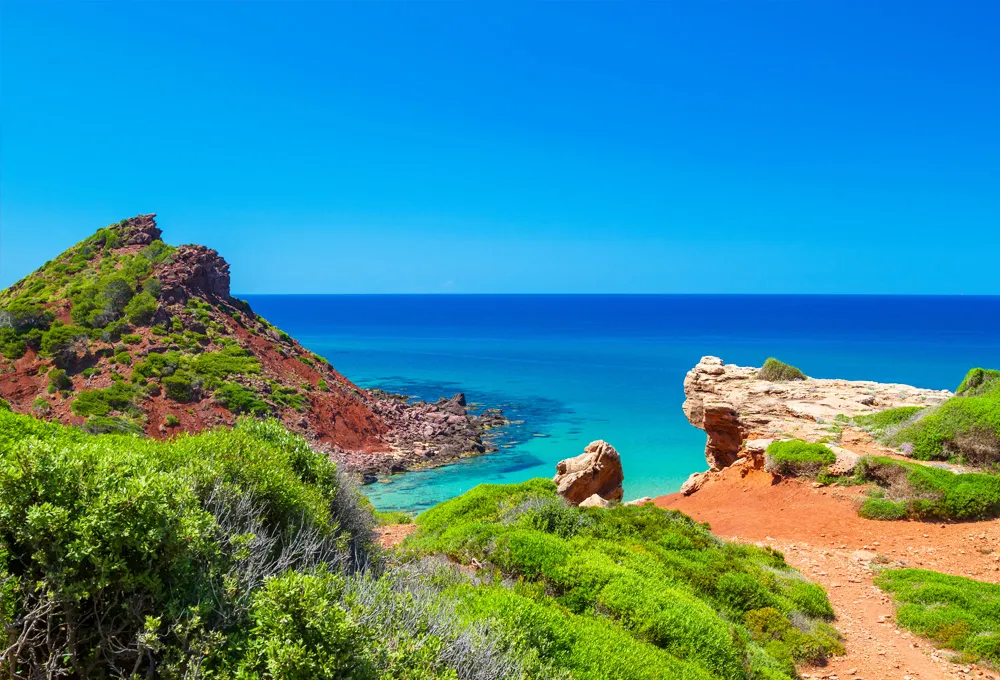 Paesaggio costiero con scogliere rosse, vegetazione verde e mare blu limpido sotto un cielo azzurro.