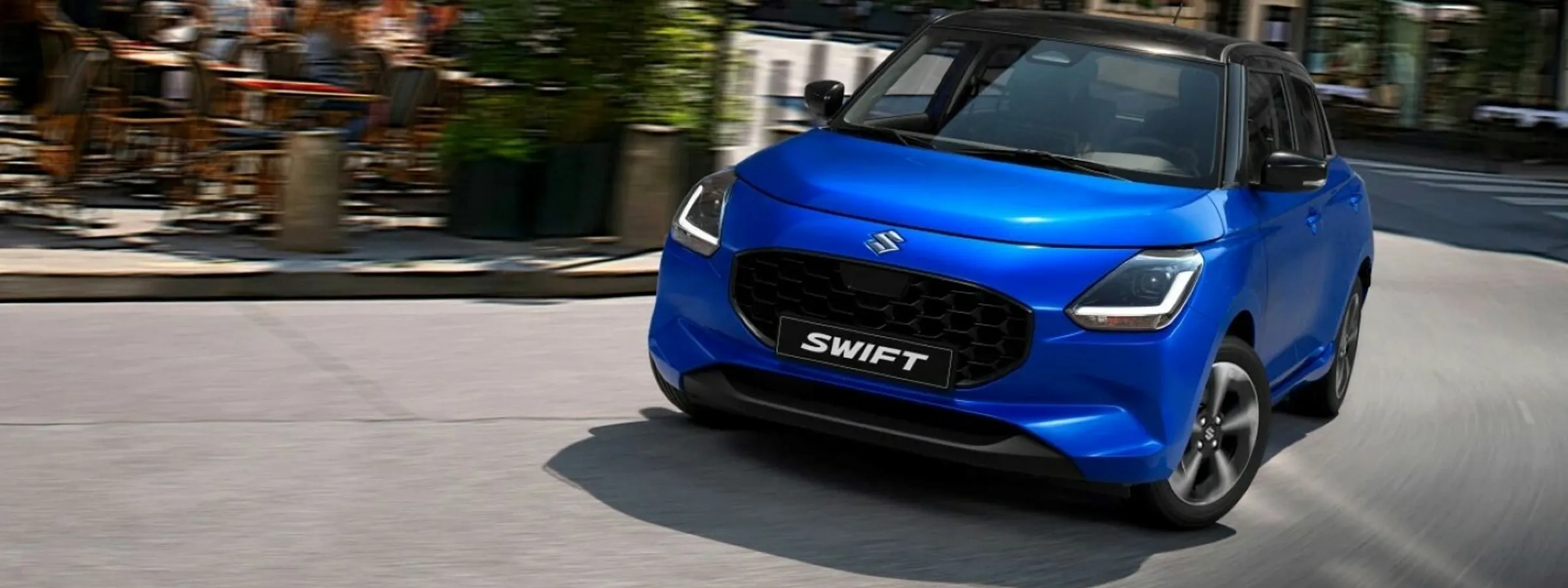 Auto a noleggio Suzuki Swift che guida su una strada urbana, blu con tetto nero.