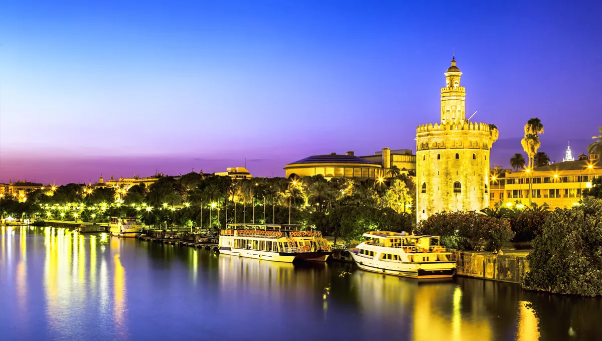 Seville river and historic Torre del Oro lit up at dusk with riverboats docked for rental cars.