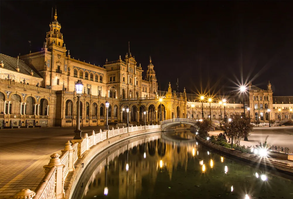 Plaza de España in Seville at night with illuminated historic buildings and reflections in the water canal.