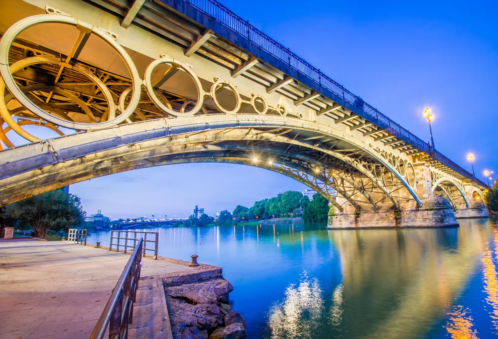 Triana Bridge in Seville illuminated at dusk over the Guadalquivir River with calm waters reflecting the lights.