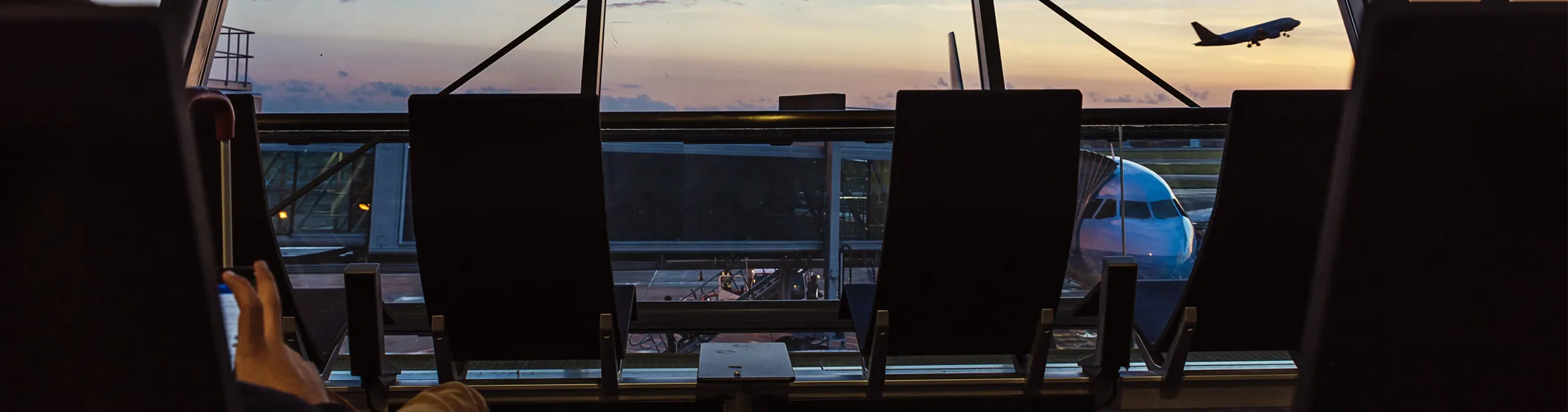 Airport terminal seating area with planes on the runway and a plane taking off at sunset.